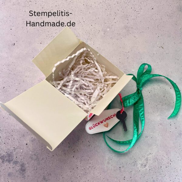 handdekorierte Fensterschachtel mit Glückwunsch-Anhänger, Geschenkbox mit Schlüsselmotiv und grünem Band, Nahaufnahme Fenster der Schachtel mit Füllung, runder Anhänger mit Schloss und Schlüssel, Schachtel geöffnet mit sichtbarem Inhalt, Glückwunsch-Tag an Geschenkbox, Geschenkverpackung für Einzug oder Prüfung